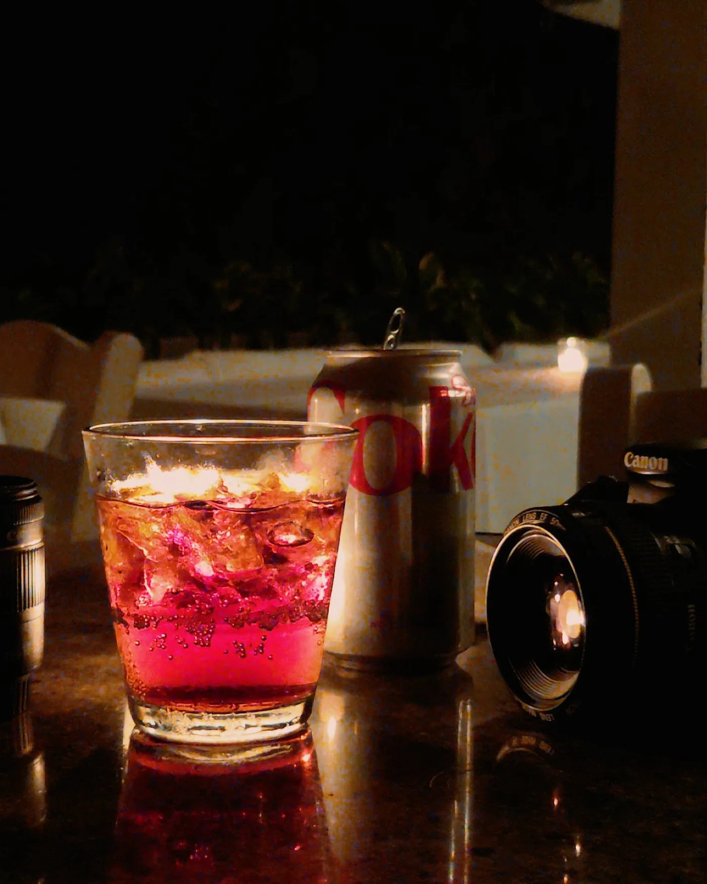 A can of cola and a glass of cola over ice on a table at night, beside a candle and a camera lens.